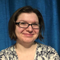 A picture of Gabrielle Steele, a white woman with glasses and chin-length dark brown hair. She is visible from the shoulders up and is in front of a blue curtain. She is facig the camera and has a slight smile.